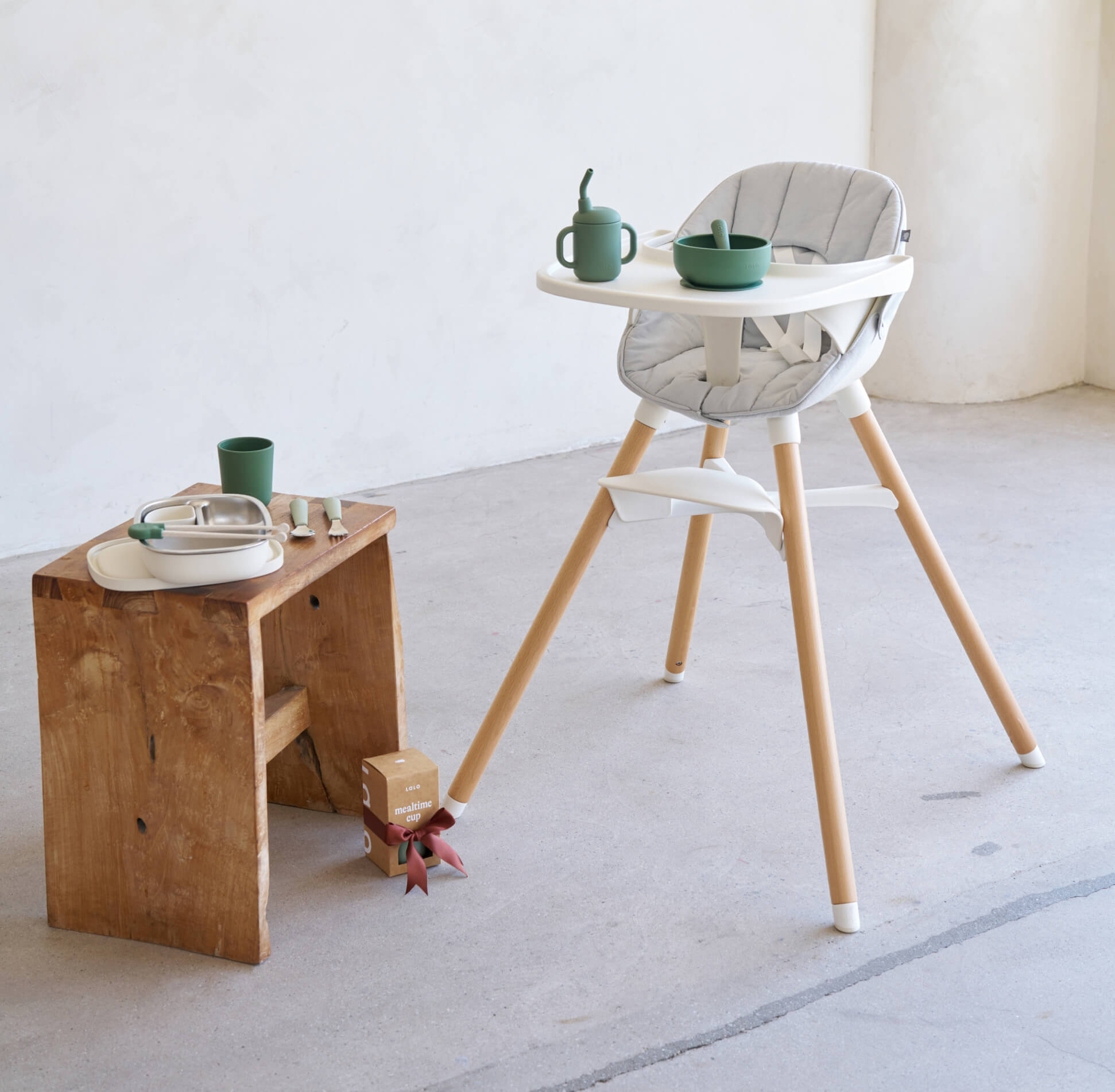 The Chair in Coconut with Olive first bites starter kit on it's tray and bento box, mealtime cup and utensils arranged on a stool next to it in an empty room